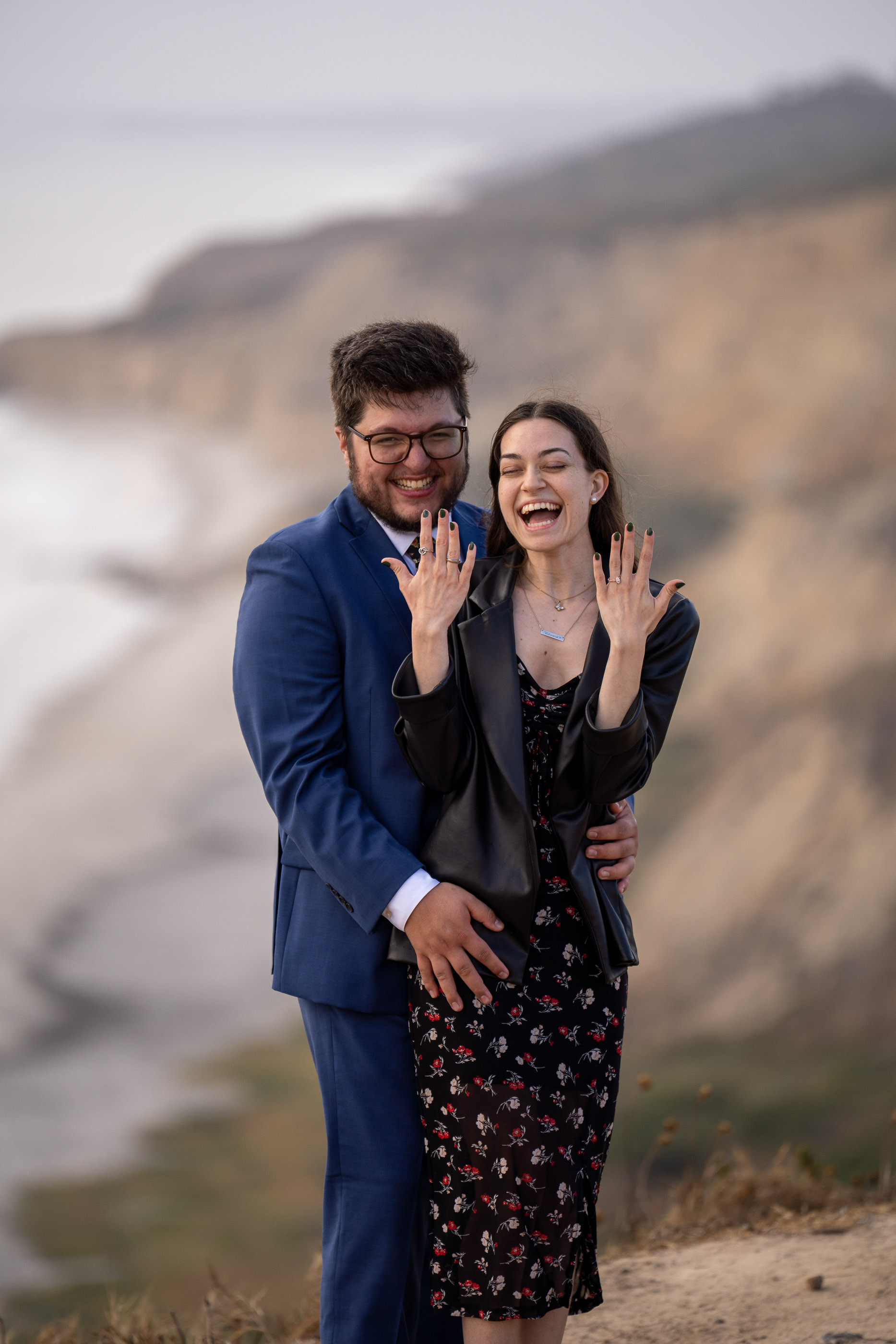 Engaged couple embracing and smiling together with a coastal landscape behind them