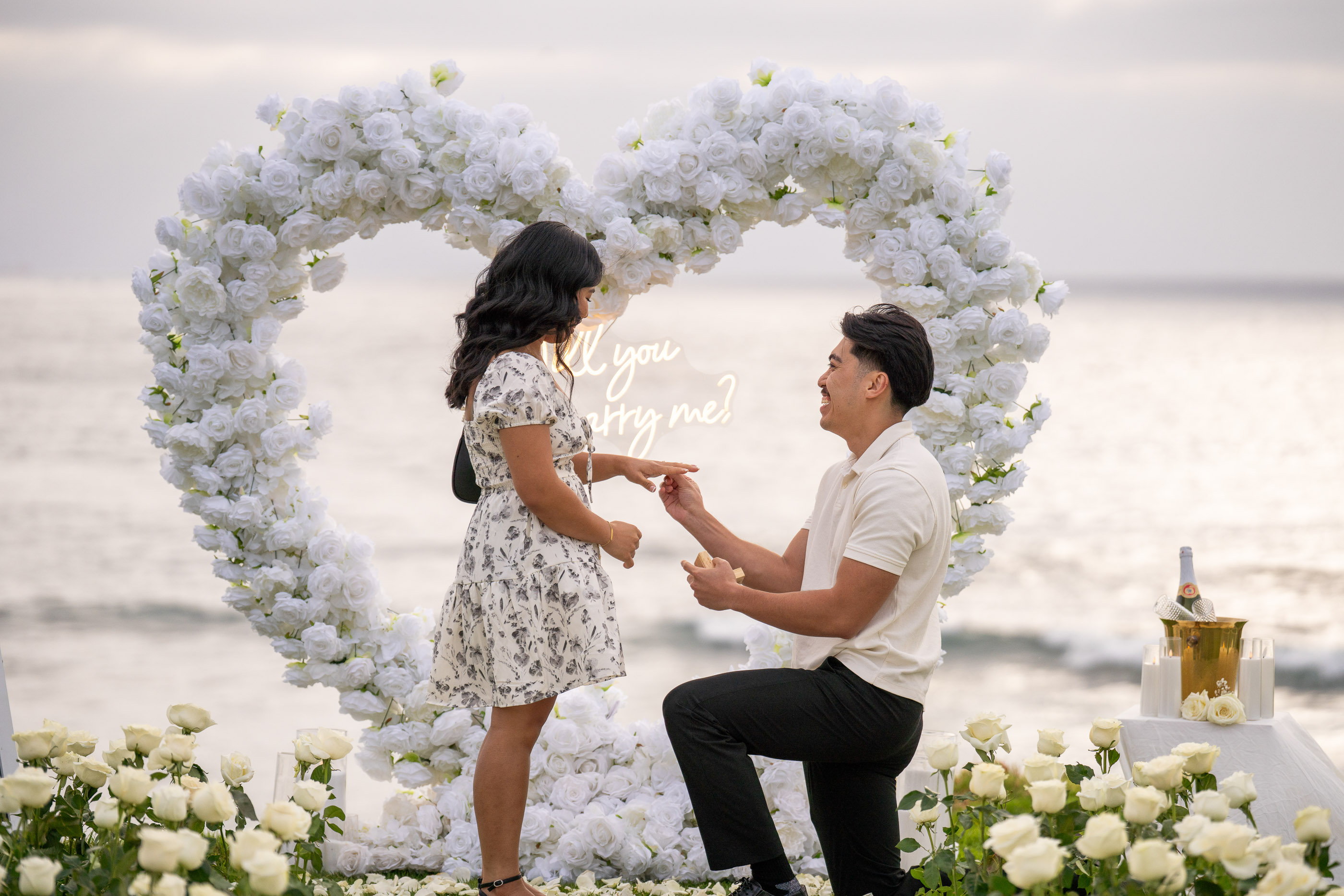 Couple sharing a proposal beneath a large white floral heart overlooking the ocean