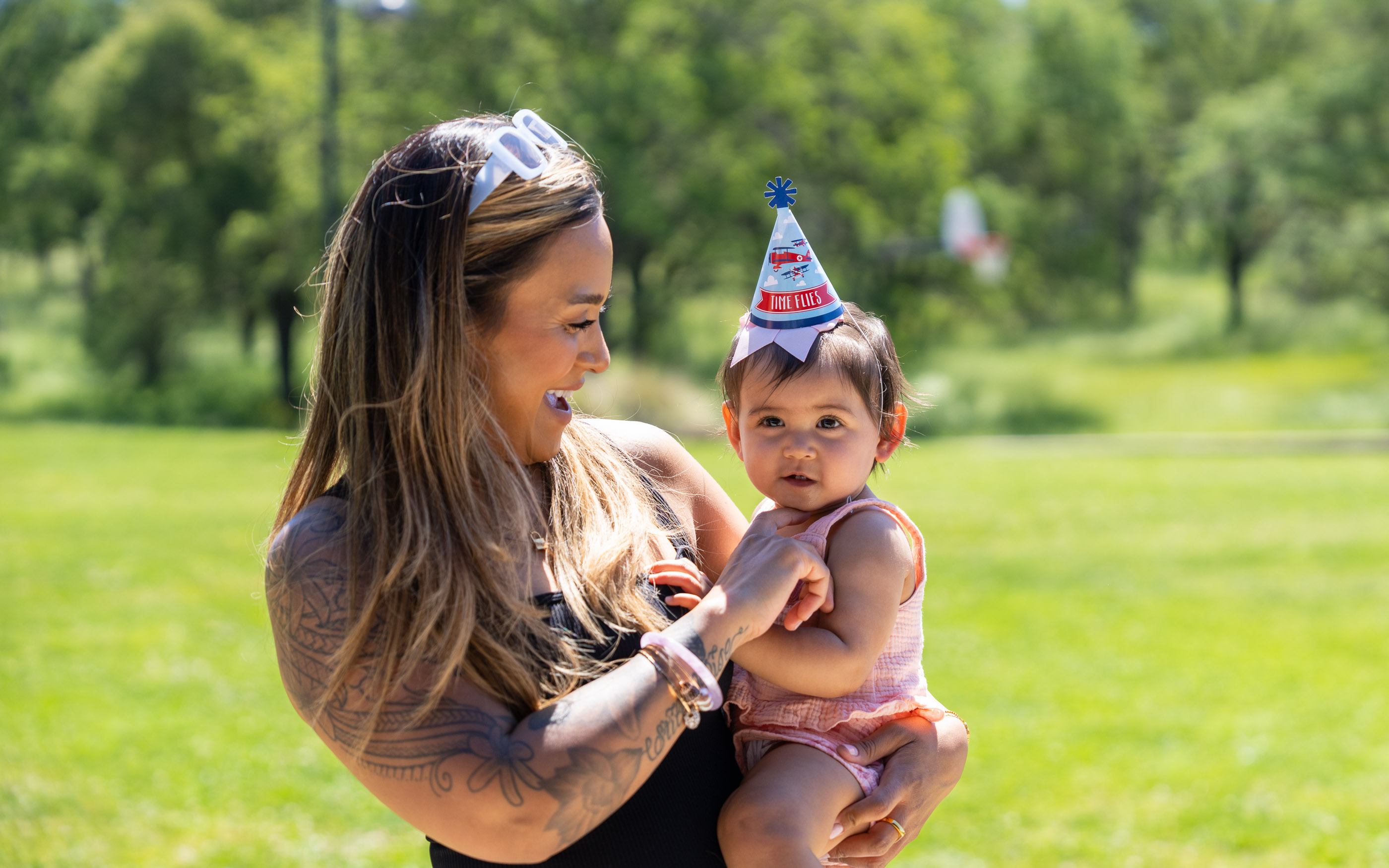 Mother holding her baby wearing a birthday hat outdoors in a sunny park