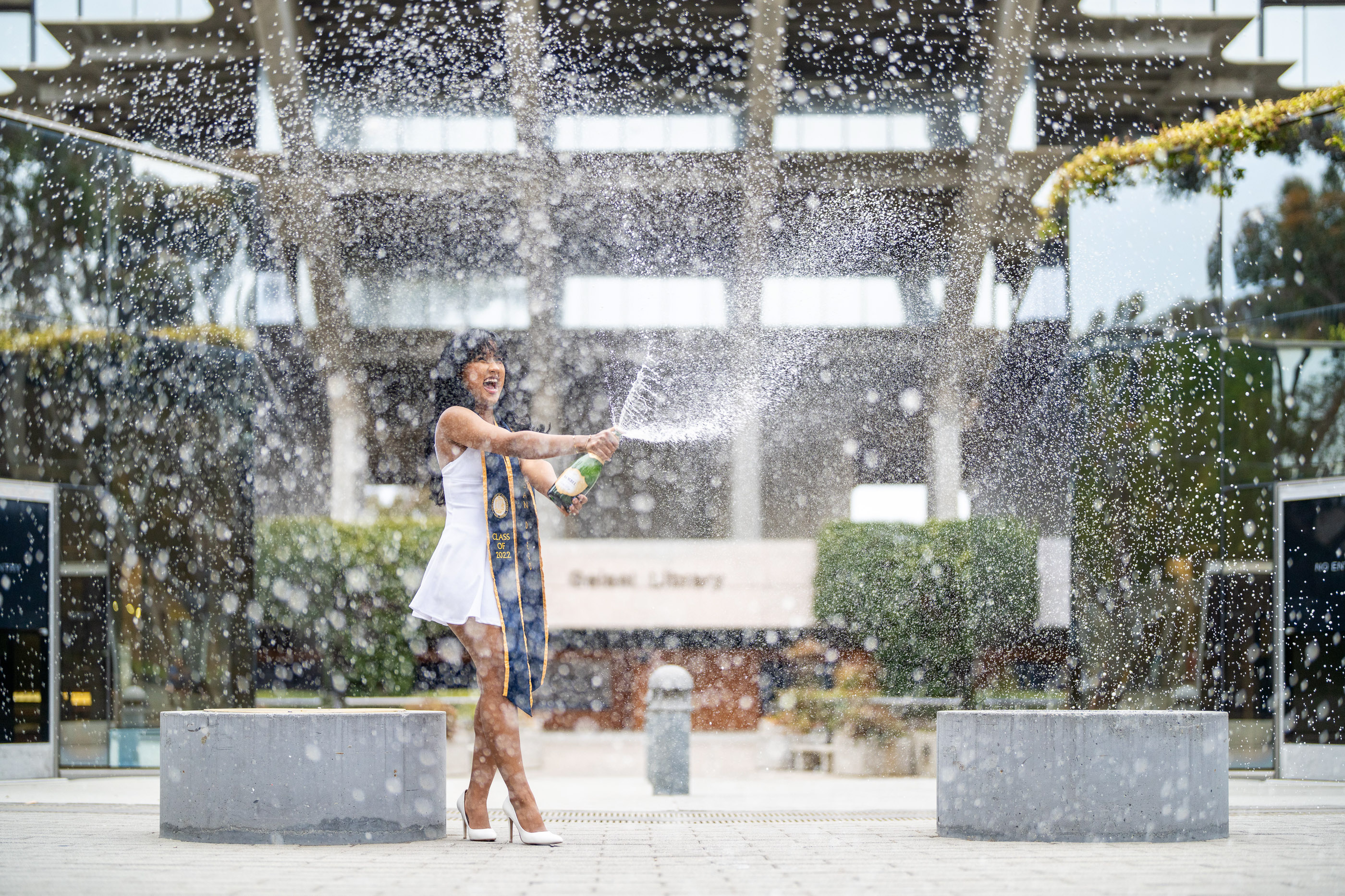 Graduate celebrating outdoors beneath a burst of falling water in a graduation gown