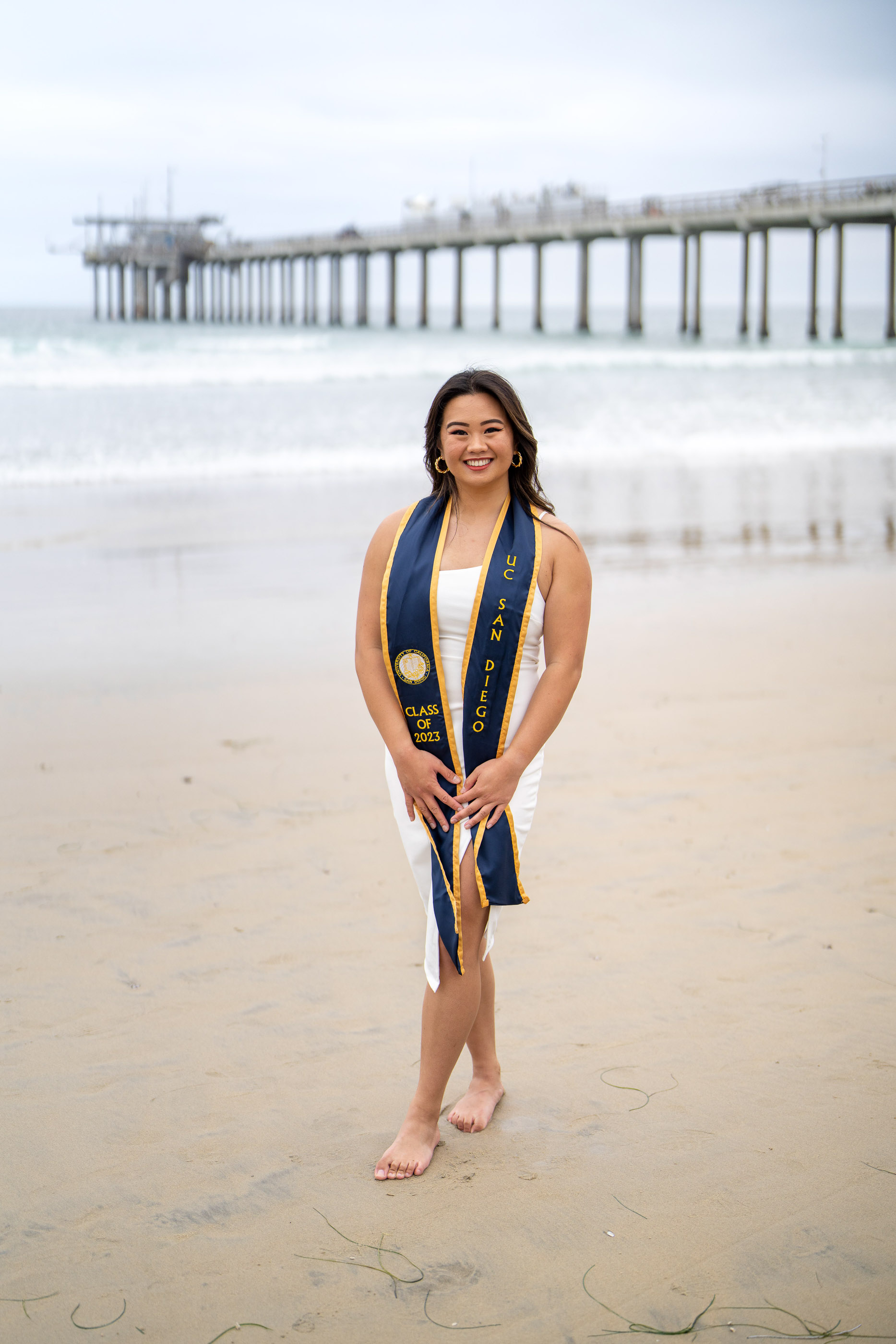 Graduate standing barefoot on the beach with a pier in the background