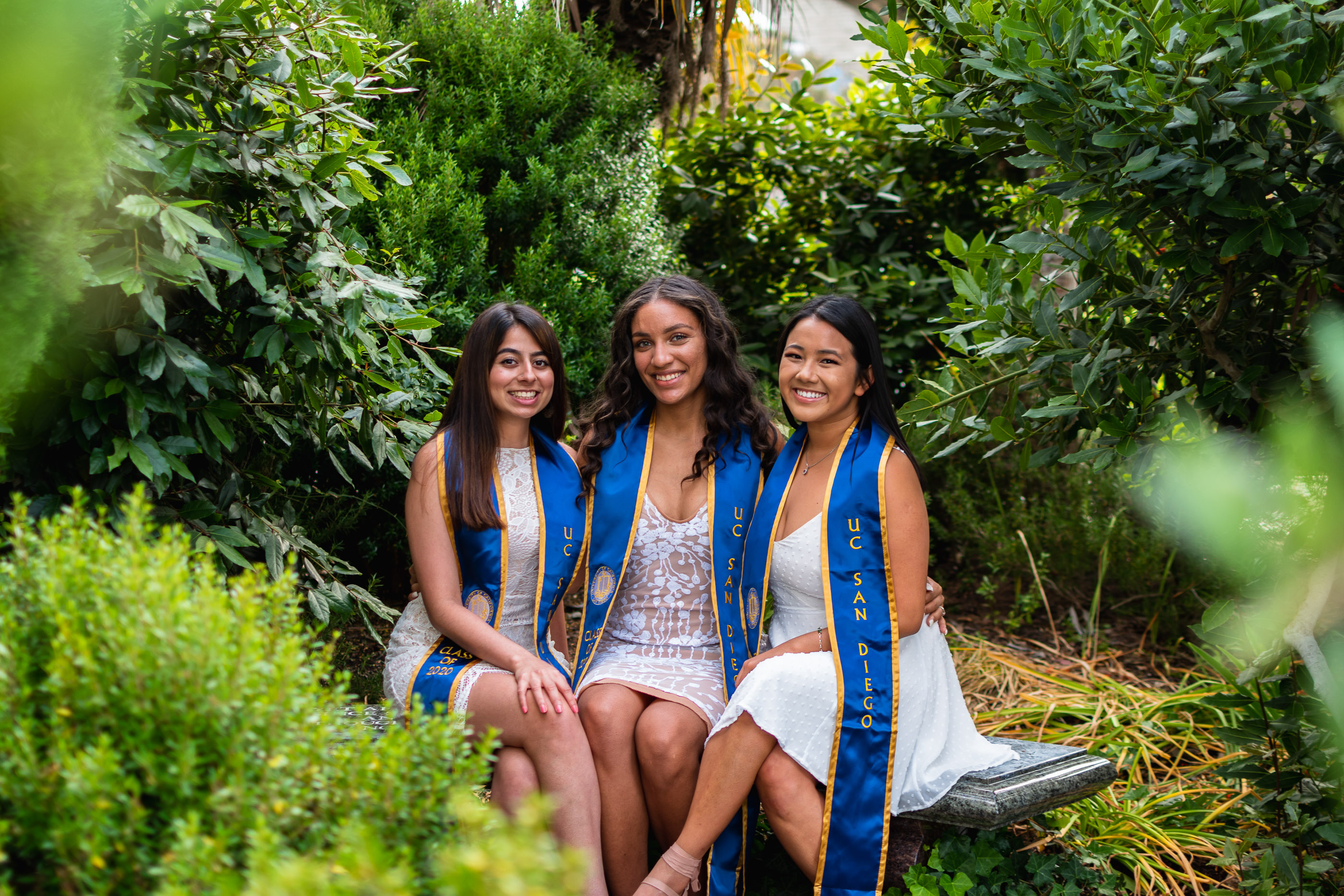 Three graduates sitting together and smiling outdoors surrounded by greenery