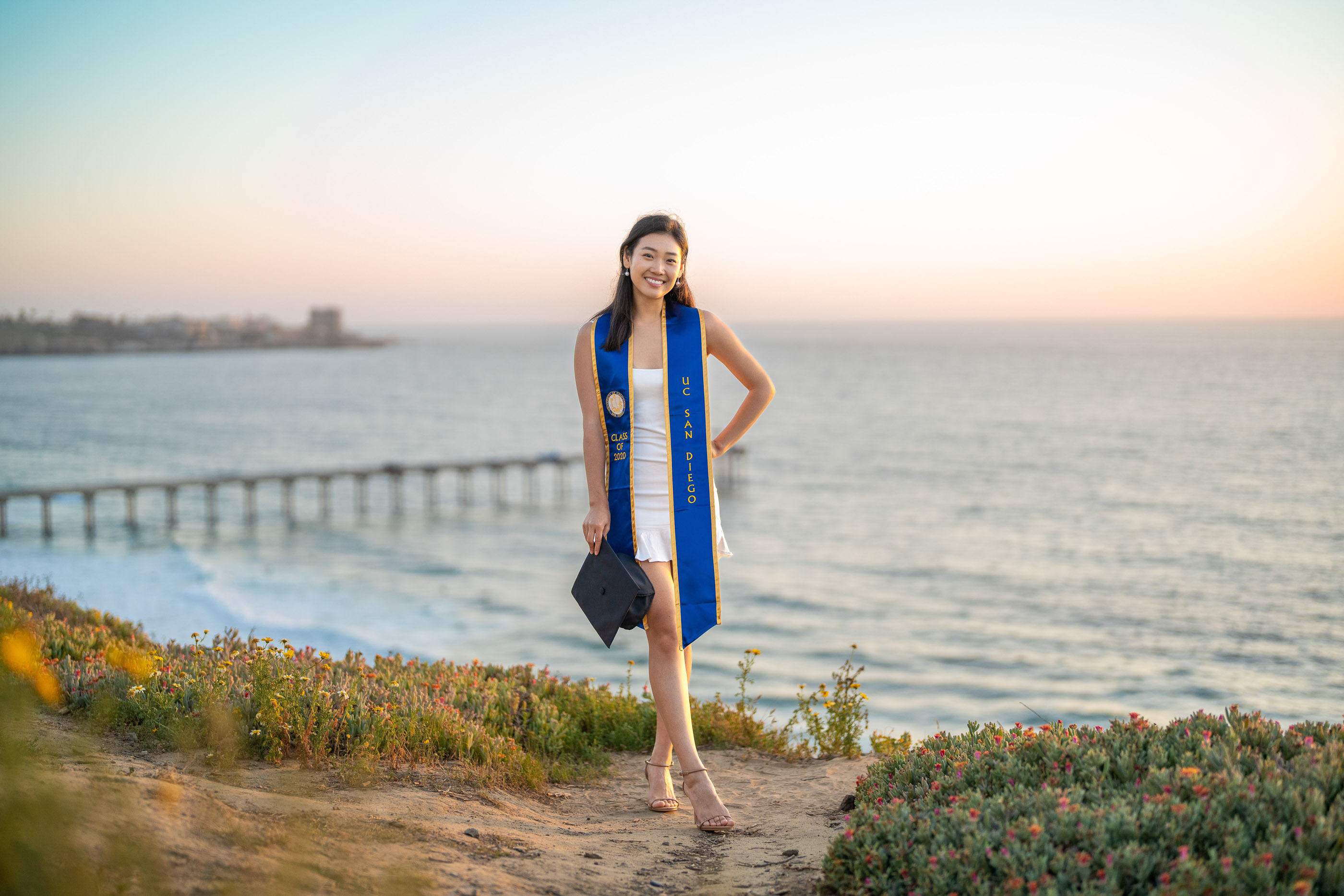 Graduate posing in cap and gown on a coastal bluff at sunset