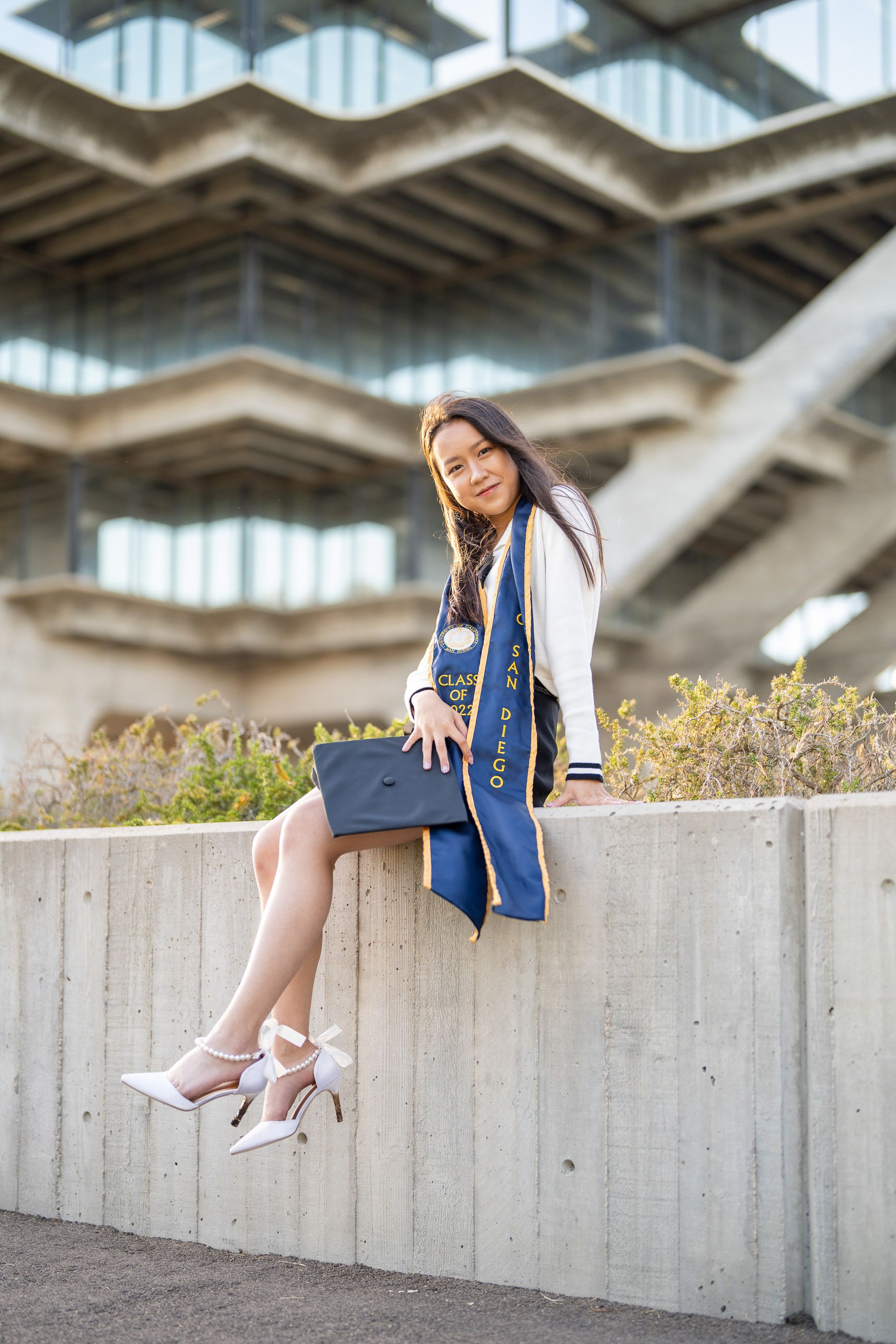 Graduate sitting on a concrete ledge in cap and gown on campus