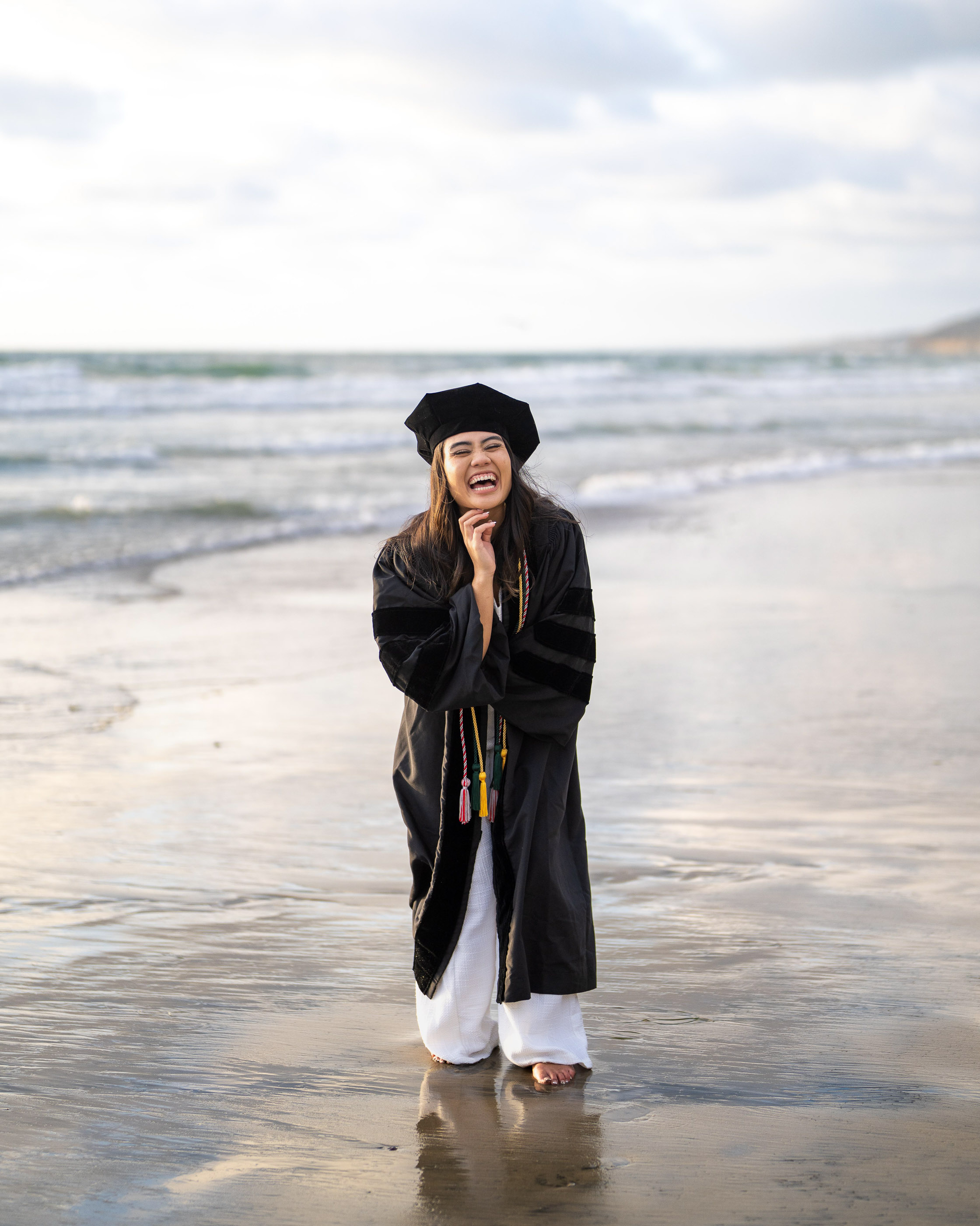 Graduate standing on the beach in cap and gown with waves behind her