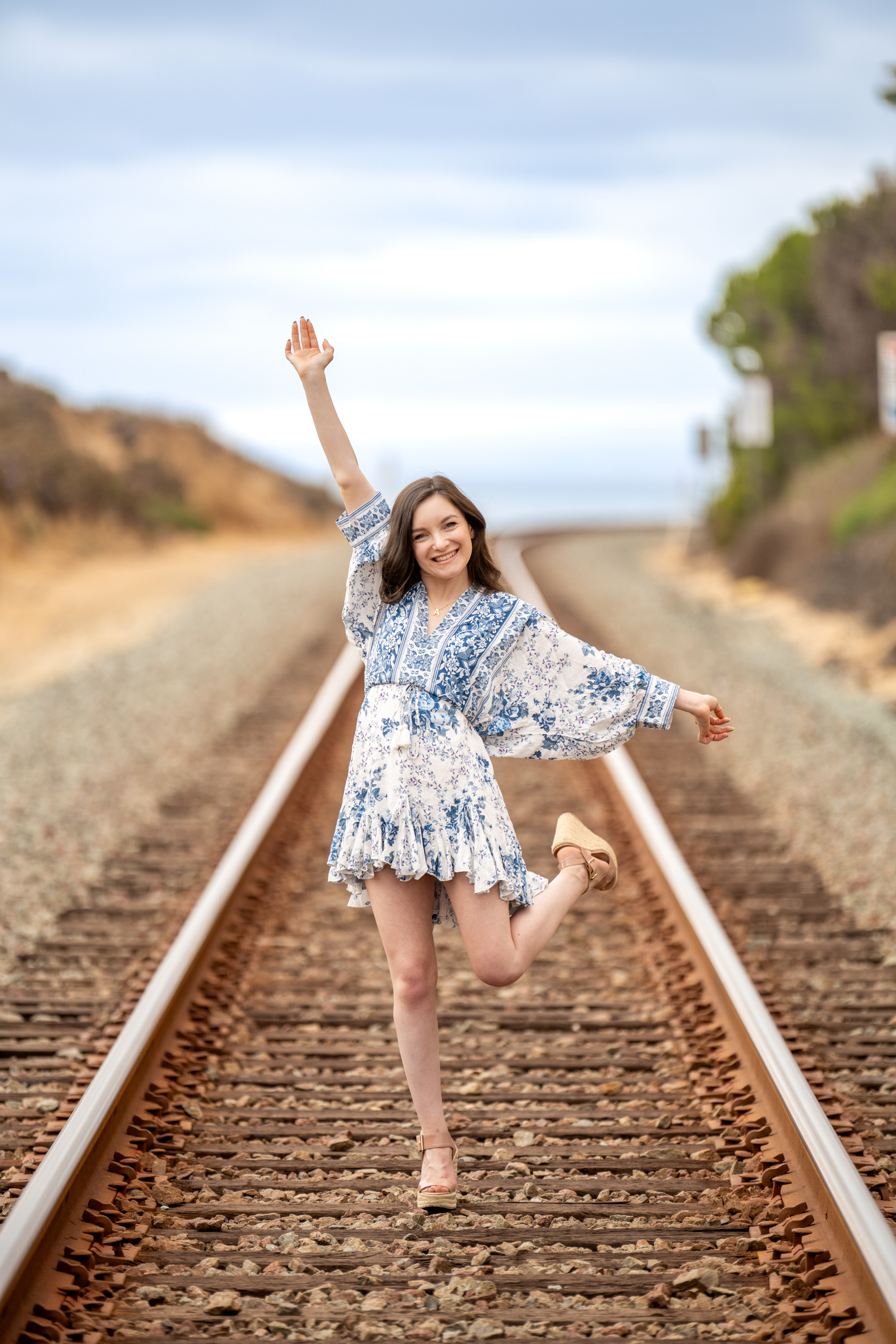 Woman happily posing on railroad tracks near Del Mar with one arm raised and one leg lifted.