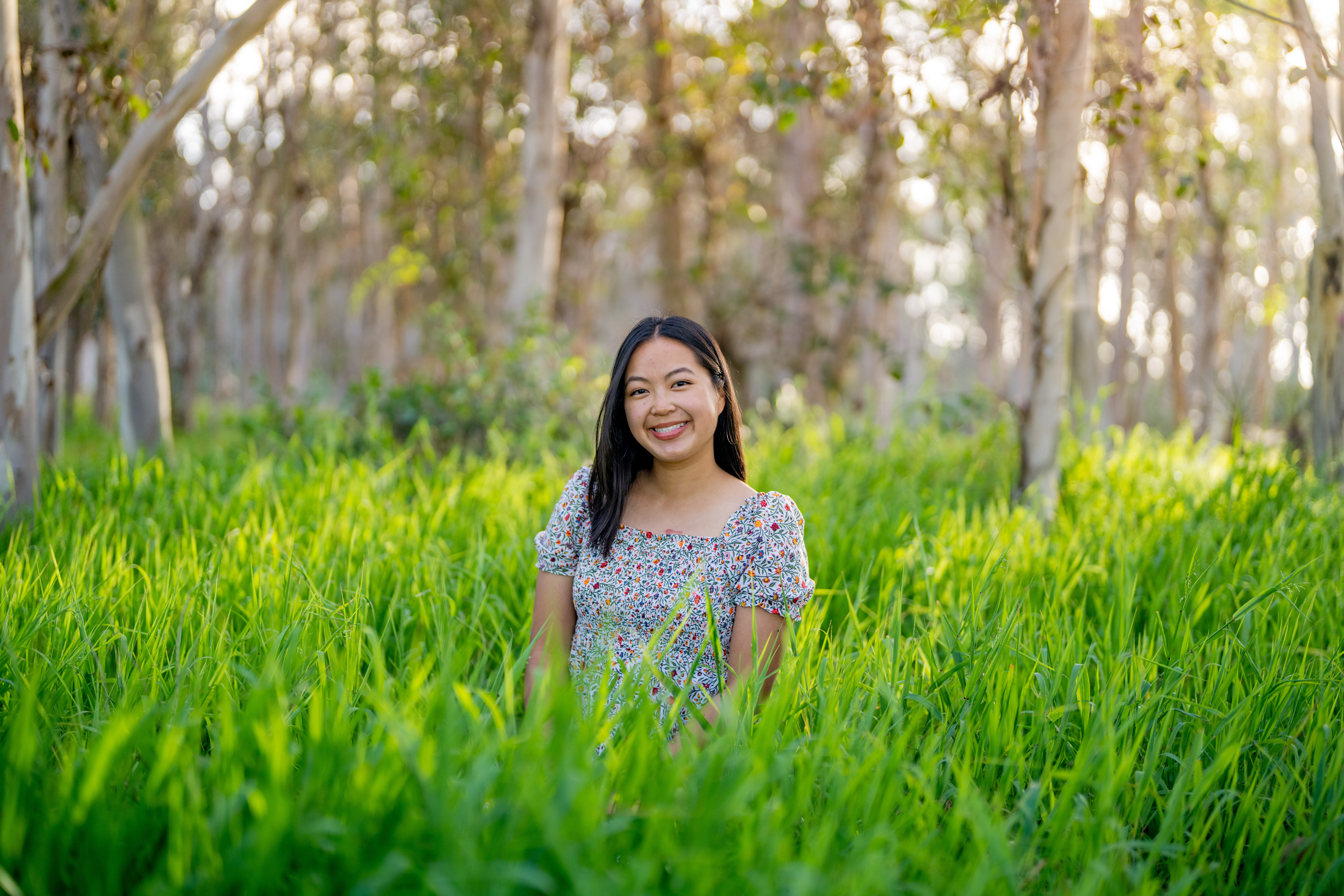 Woman smiling for a portrait while standing in tall green grass among eucalyptus trees.