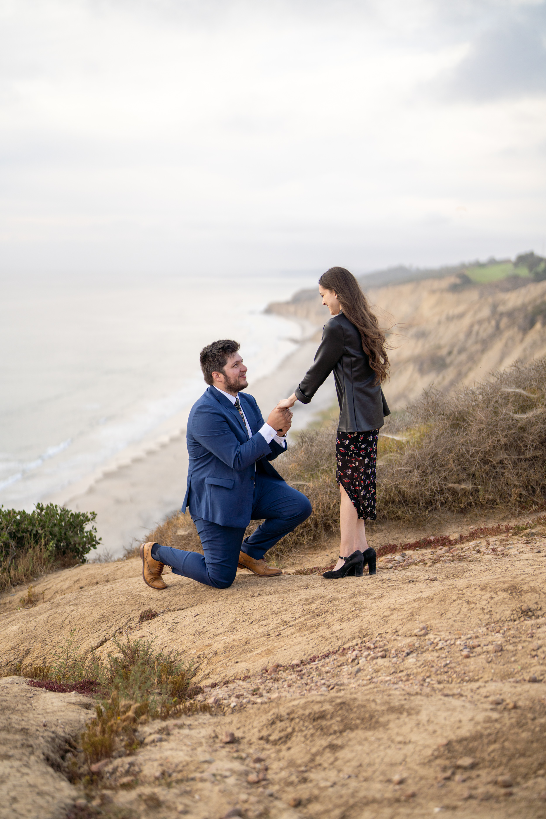 Couple at the coastal cliffs near Gliderport as a man kneels to propose to a woman.