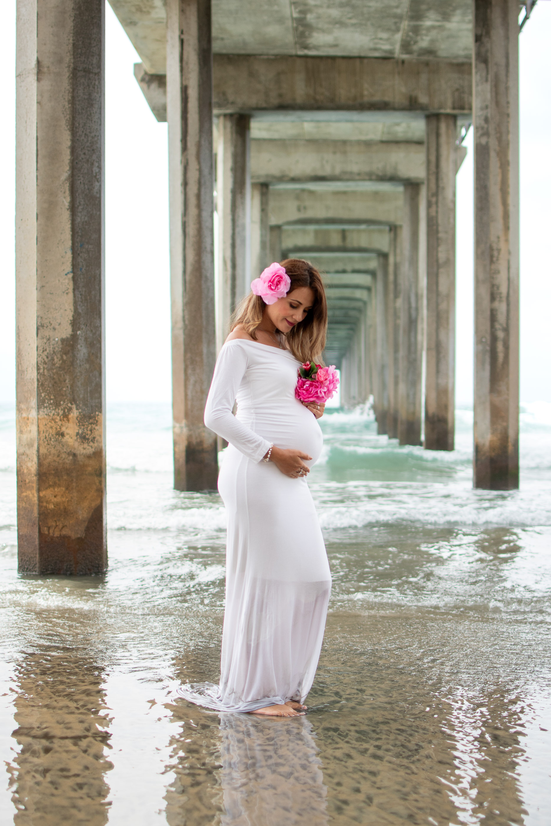 Pregnant woman walking beneath a covered beach walkway in a flowing white dress